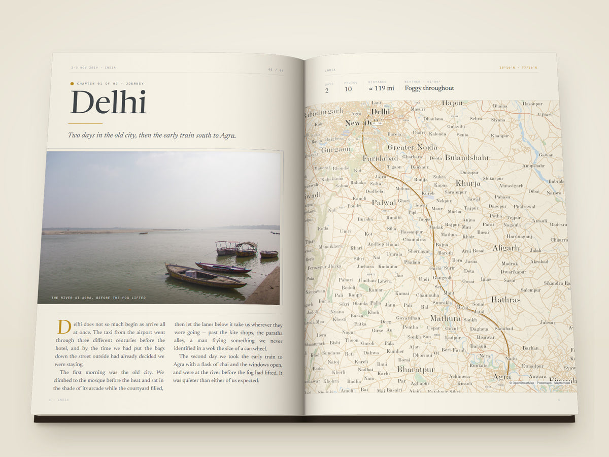 The same A4 portrait softcover Travel Book, open on the table. Two more printed pages — A chapter opening set like a magazine feature — Delhi large on the left page over a photograph of boats on the river at dawn, an italic line and a two-column essay with a drop capital, facing a full-page map of the road from Delhi to Agra with the chapter's days, photographs, distance and weather above it