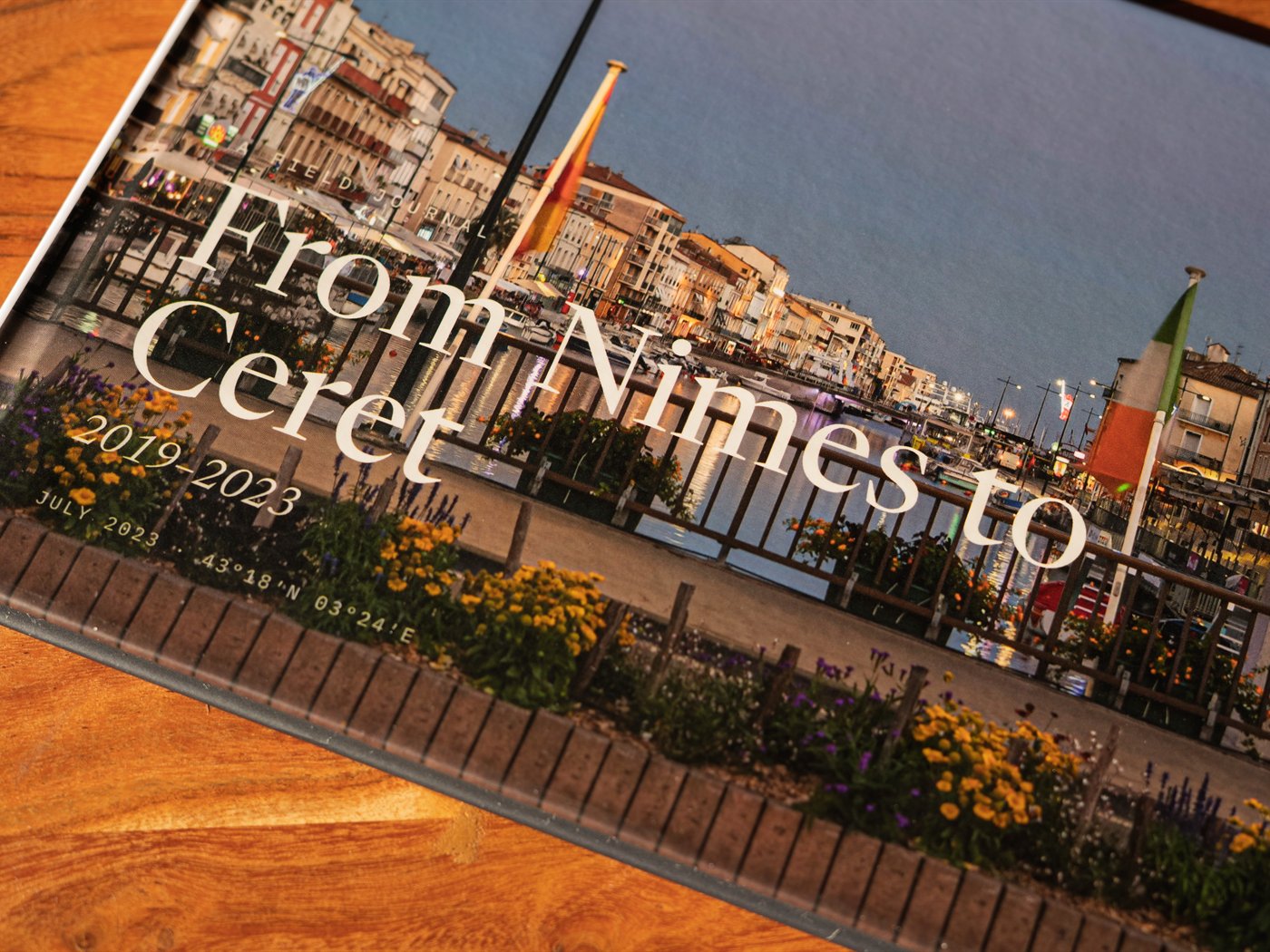 The cover of From Nîmes to Céret, close on a walnut table — the title set over a harbour at dusk