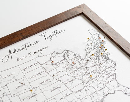 Close-up of framed 11x14" push pin USA map in black and white with metallic pins (gold, rose gold, silver, and dark silver) marking locations. Compact desk-sized map with labeled U.S. states and interstate highways.