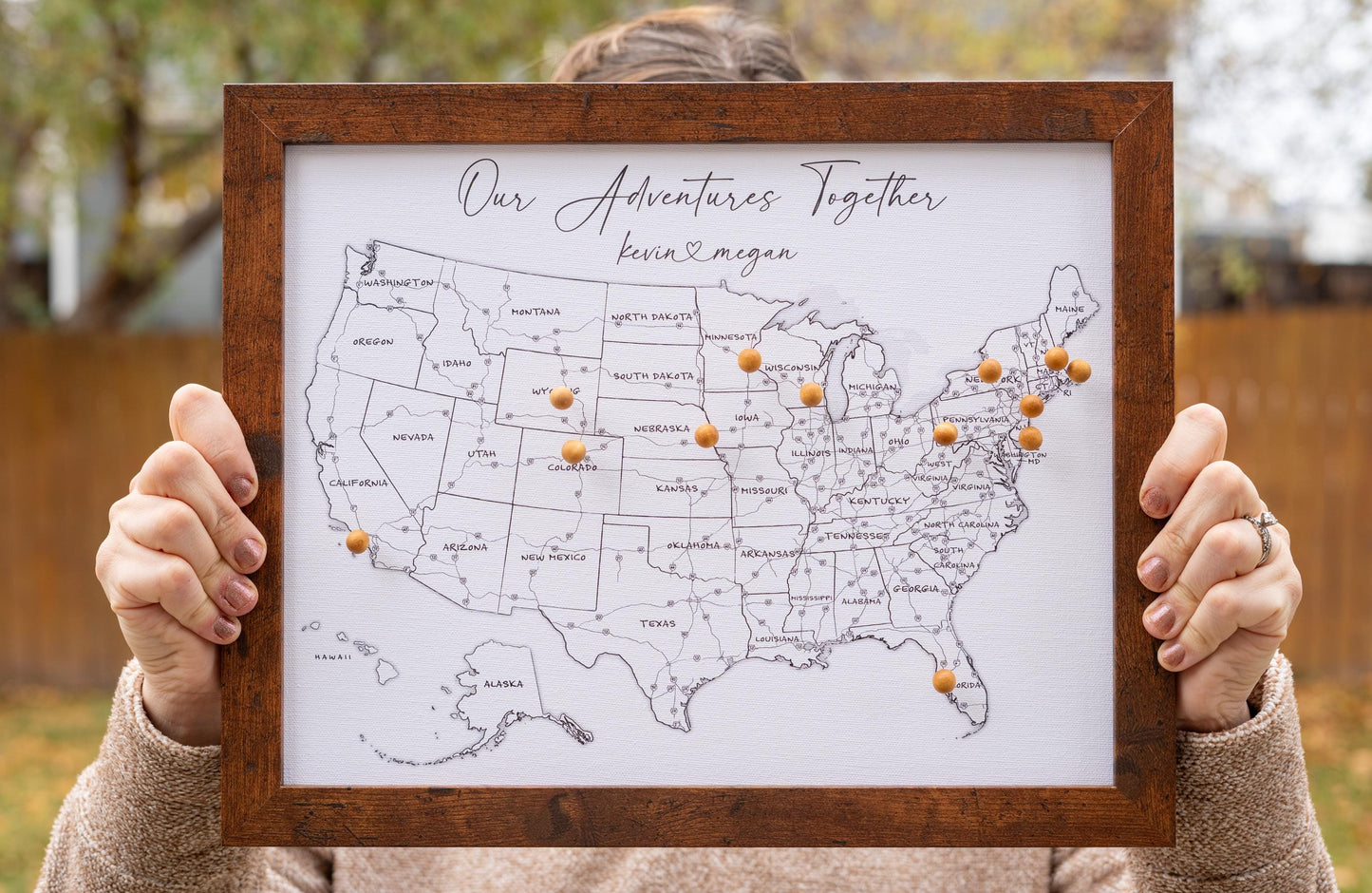 Woman holding 11x14" framed USA push pin map with wooden pins, facing camera for size reference. Black-and-white design with labeled states and highways, ideal for tracking travels and personalizing