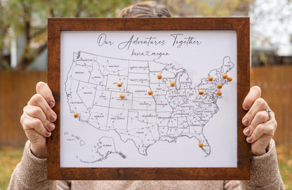 Woman holding 11x14" framed USA push pin map with wooden pins, facing camera for size reference. Black-and-white design with labeled states and highways, ideal for tracking travels and personalizing