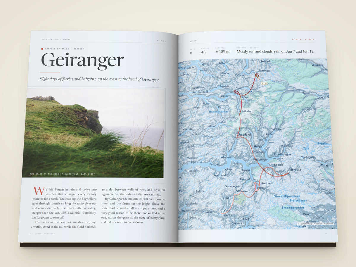 The same A4 portrait hardcover Travel Book, open on the table. Two printed pages — A chapter opening set like a magazine feature — the chapter name large on the left page over a photograph of green cliffs above the sea, an italic line under it and a long essay in two columns with a red drop capital, facing a full-page map of the fjords with the days, photographs, distance and weather set above it