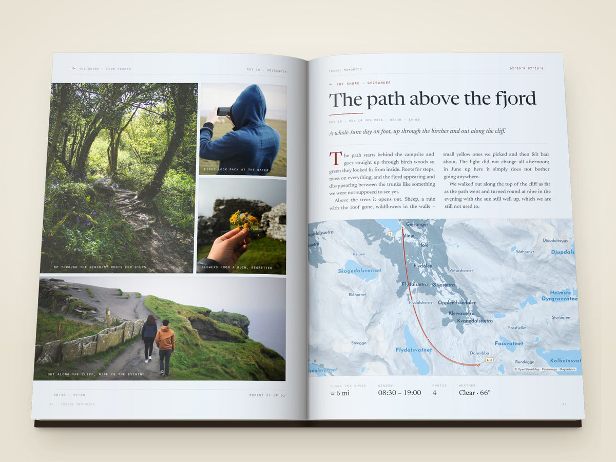 The same A4 portrait hardcover Travel Book, open on the table. Two more printed pages — A moment page — four photographs of a forest path, a phone held up to a field, wildflowers and two walkers on a cliff edge, each with its own caption, facing the title, a written account of the day, a map of the path above the fjord and the day's numbers