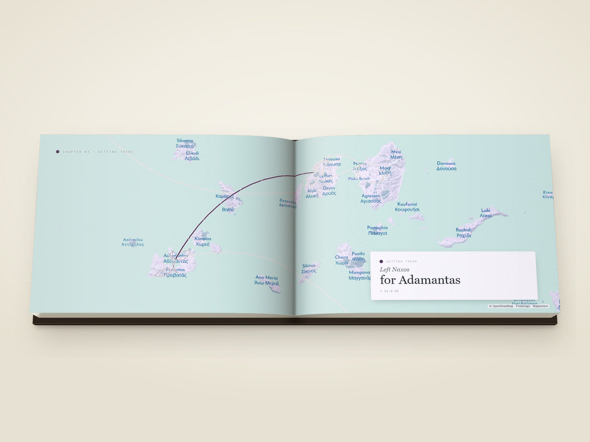 The same A5 landscape softcover Travel Book, open on the table. A getting-there spread — the leg from Naxos to Adamantas drawn as a single arc across a map filling both printed pages, with a card noting the departure